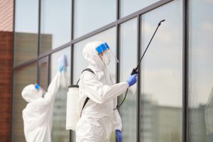 Portrait of two workers wearing protective suits spraying chemicals over building outdoors during disinfection or cleaning, copy space